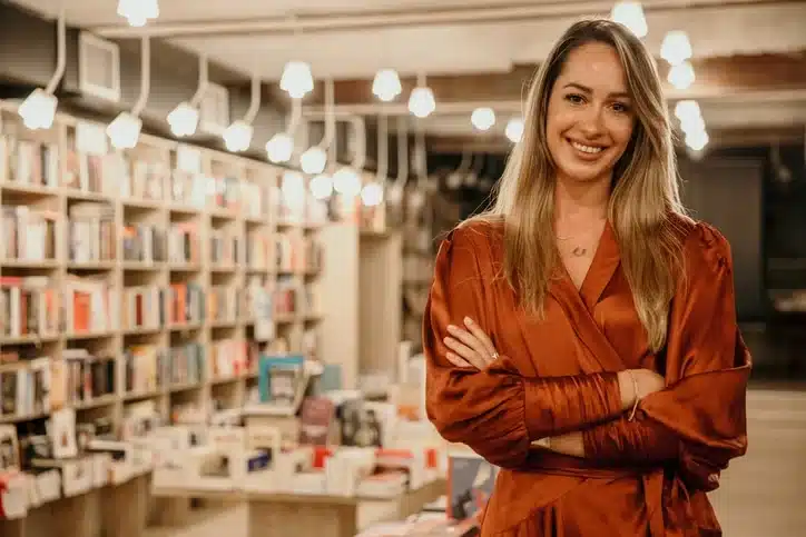 A smiling woman in a burnt orange blouse standing with crossed arms in a bookstore with shelves of books in the background and overhead lighting.