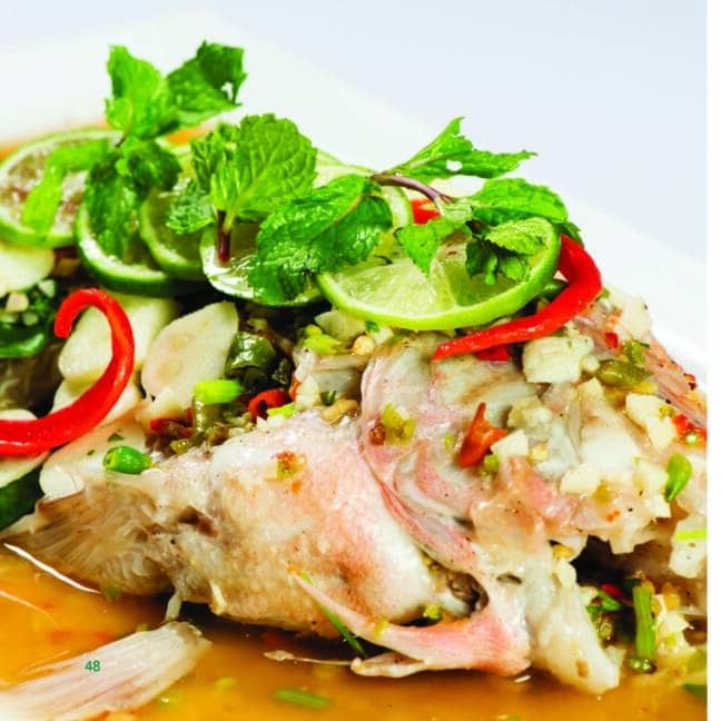 A steamed whole fish topped with fresh herbs, slices of lime, red chili peppers, and diced green onions, served in a light, savory sauce.