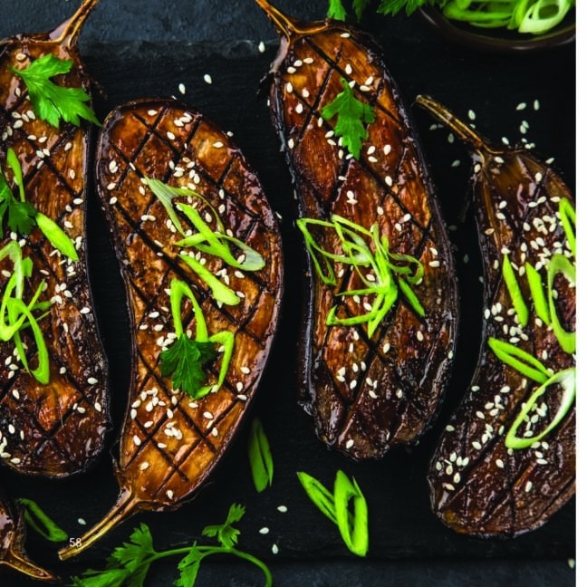 Grilled eggplant halves garnished with sesame seeds, green onion strips, and parsley on a dark slate surface.