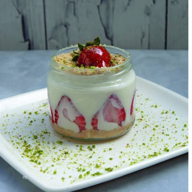 A dessert in a glass jar, featuring layers of crumbled biscuits, creamy filling, and fresh strawberry slices, topped with a whole strawberry and garnished with crushed pistachios, served on a white plate dusted with green pistachio crumbs.